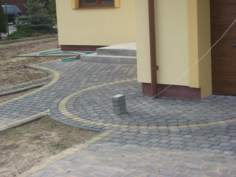 Ułożona kostka brukowa w szarych odcieniach z żółtymi obrzeżami tworząca ścieżkę w kształcie łuku przed budynkiem. Na bruku leży stos kostki, a obok widać przygotowane podłoże.